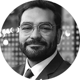 Smiling man with glasses and a beard in a suit, looking at the camera with a blurred city background.