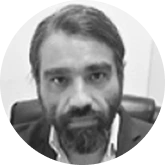 Black-and-white portrait of a man with dark hair and a full beard, looking at the camera while seated in an office chair, wearing a collared shirt and jacket.
