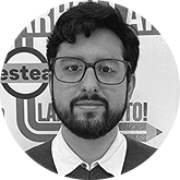 Portrait of a bearded man with glasses, wearing a white shirt and tie, posed in front of a backdrop with circular logos.