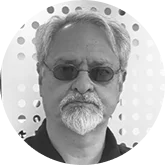 Grayscale close-up portrait of an older man with gray hair, a goatee, and dark sunglasses facing forward with a dotted background.