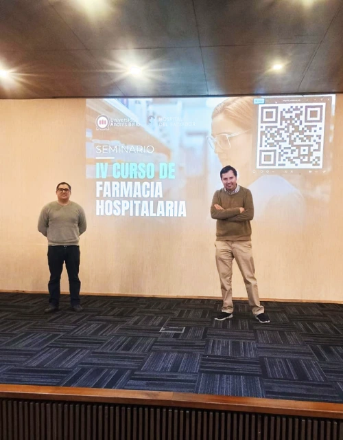 Two men stand on a stage in front of a projected slide that reads 'IV CURSO DE FARMACIA HOSPITALARIA' with a QR code in the top-right corner.