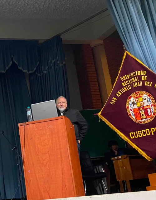 Bearded man giving a talk at a wooden podium on a stage, with a laptop and water bottle, blue curtains, and a maroon pennant with a crest visible beside him.