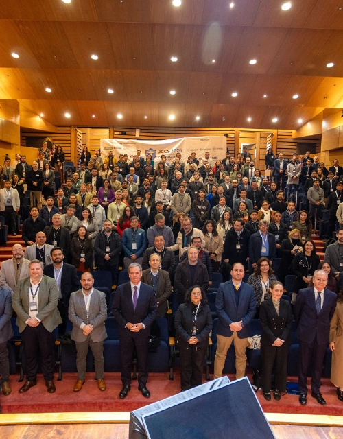 Large group of conference attendees posing on a stage and in rows inside a wood-paneled auditorium.
