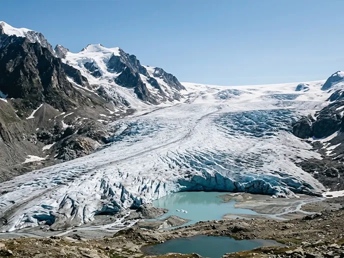 Glacier valley with rugged snow‑capped mountains, a blue glacier front, and small turquoise meltwater pools in the foreground under a clear sky.